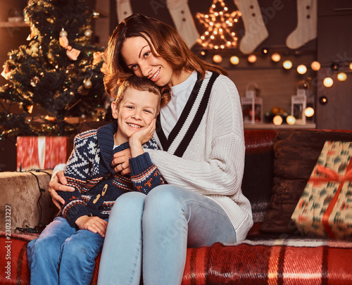 Mom hugging her cute little boy while sitting on sofa in decorated room during Christmas time.