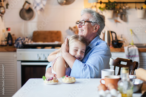 Happy grandmother is hugging granddaughter in cozy home kitchen. Family is cooking together. Senior woman and cute little child girl are smiling. Kid is enjoying kindness, warm hands, care, support. photo