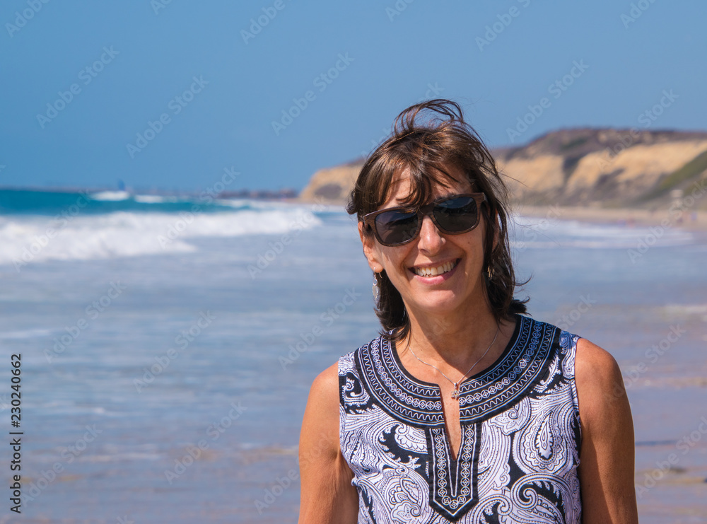 Portrait view of happy baby boomer woman smiling at the beach with ocean water and large cliff if mountain in background. Portrait view of happy baby boomer woman smiling at the beach with ocean water and large cliff if mountain in background.