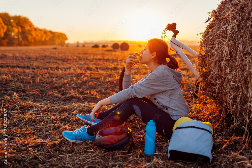 Naklejka premium Young bicyclist having rest after a ride in autumn field at sunset. Woman eating by haystack