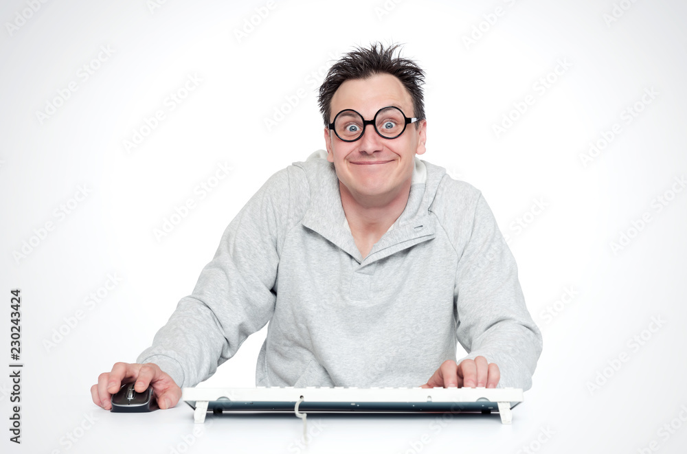 Happy smiling man in round glasses sits at a table with a keyboard and mouse in front of a computer. Satisfied programmer Happy smiling man in round glasses sits at a table with a keyboard and mouse in front of a computer. Satisfied programmer