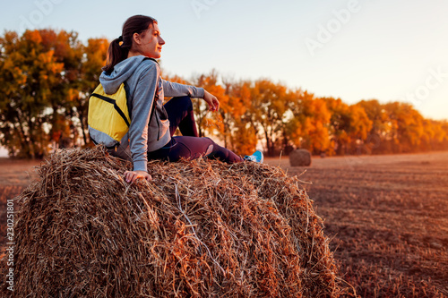 Young traveler having rest in autumn field at sunset. Woman admiring view sitting on haystack