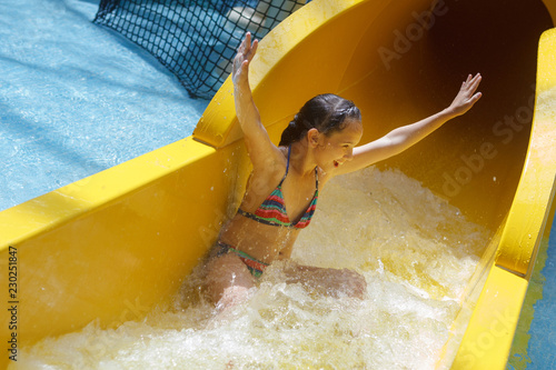 Young pretty girl in a striped colorful swimsuit laughs, rides on a yellow water slide in aqua park.