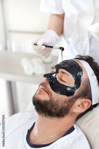 Kosmetyczka nakłada mężczyźnie maseczkę na twarz w salonie kosmetycznym.