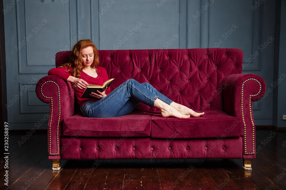 Rich young redhead woman reading a book on red velvet sofa. Rich young redhead woman reading a book on red velvet sofa.