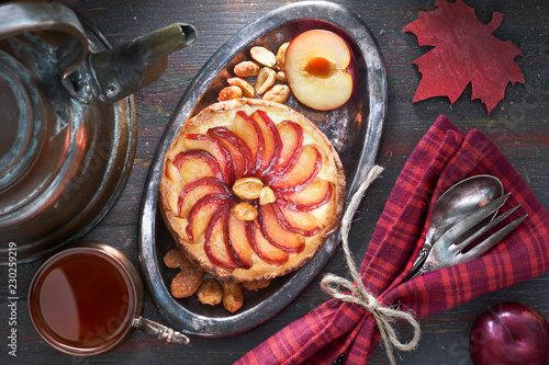 Homemade plum tart with peanuts on metal plate with crockery, black tea and tea kettle on dark wood