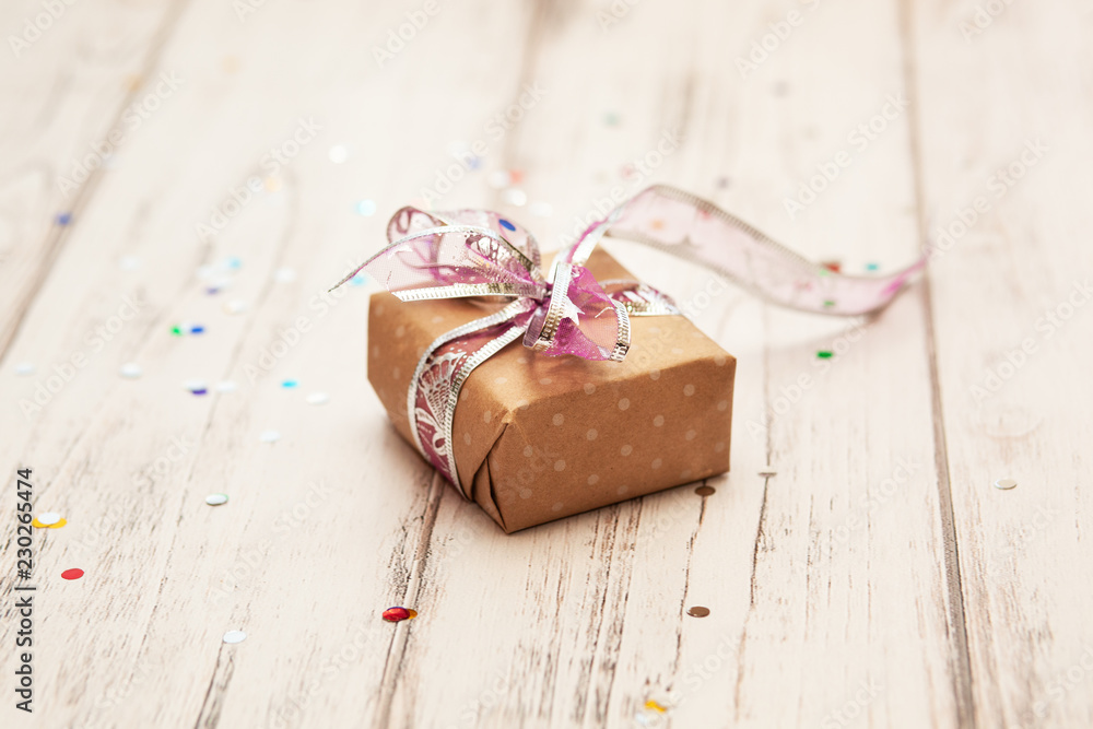 brown gift box with ribbon on white wooden board background with confetti. Selective focus brown gift box with ribbon on white wooden board background with confetti. Selective focus