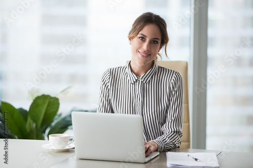 Head shot portrait of confident businesswoman at workplace, smiling woman employee sitting behind laptop and looking at camera. Staff at work. photo