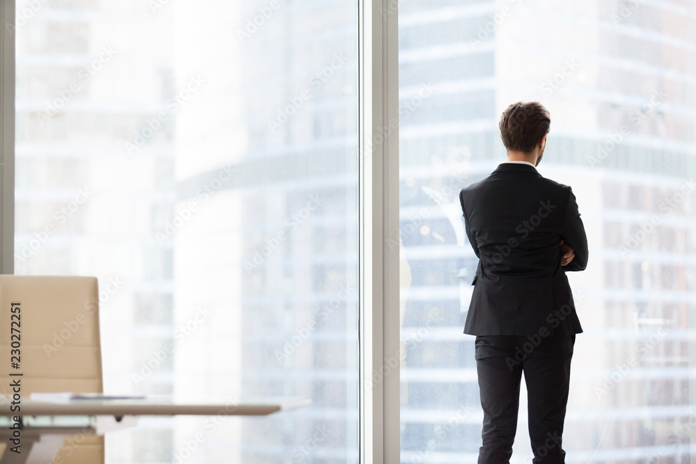 Back view portrait of successful confident businessman, property investor standing by window. Boss looking at city from cabinet with large windows, spacious office. Calm mind, business vision concept Back view portrait of successful confident businessman, property investor standing by window. Boss looking at city from cabinet with large windows, spacious office. Calm mind, business vision concept