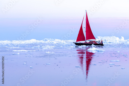 Beautiful red sailboat sailing among icebergs during dawn. Ilulissat, Greenland.