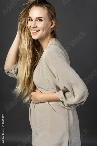 Beautiful young pregnant woman blonde, wearing a short light gray dress, happily smiles and touches her hair with her hand. Gray background. Maternity, lifestyle. Commercial design.