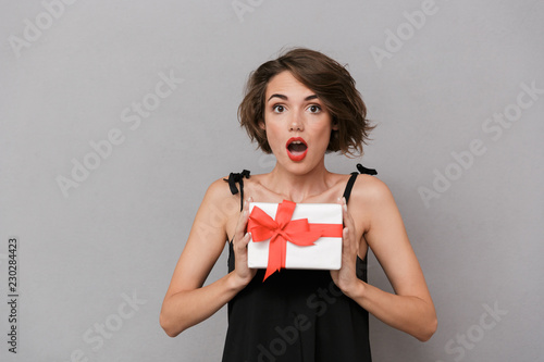 Photo of shocked woman 20s wearing black dress holding present box, isolated over gray background