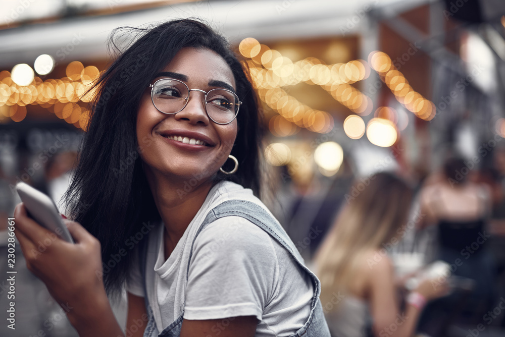 Portrait of hipster young lady in glasses holding smartphone and looking away with smile. Street with people on blurred background with bokeh effect - obrazy, fototapety, plakaty 