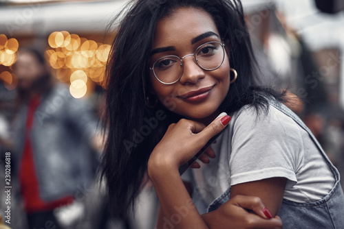 Portrait of hipster young lady in glasses holding mobile phone and looking at camera with smile. Street and people on blurred background with bokeh effect