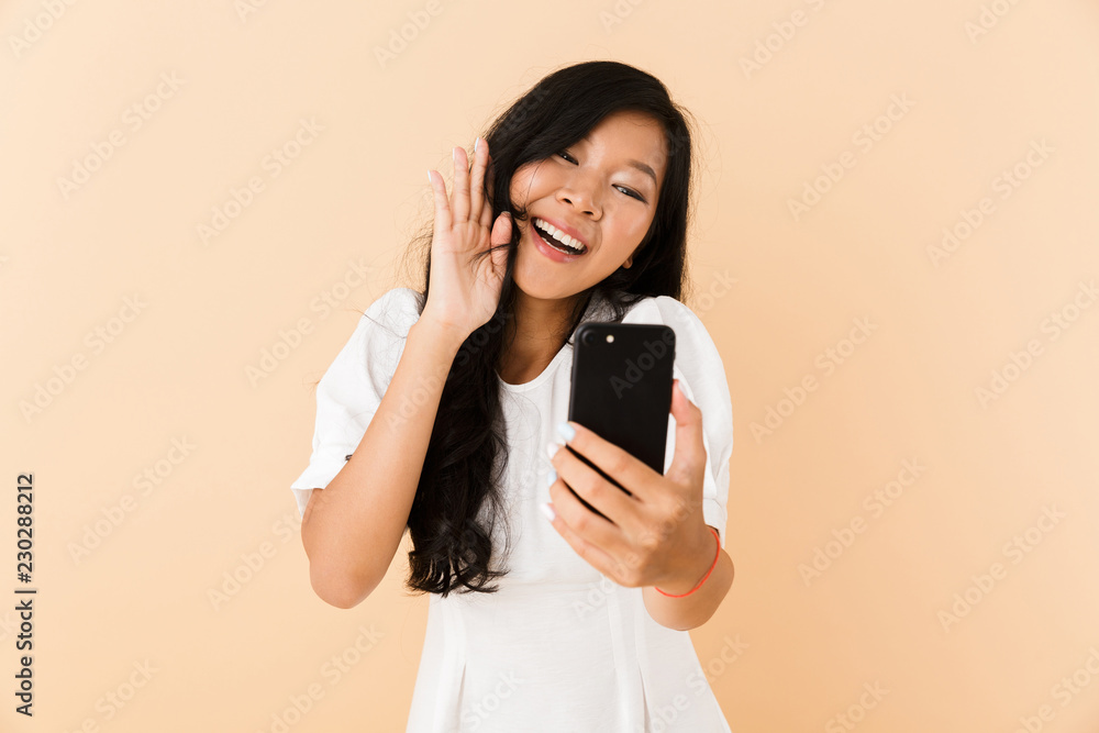 Happy asian woman posing isolated over beige wall background waving talking by phone. Happy asian woman posing isolated over beige wall background waving talking by phone.