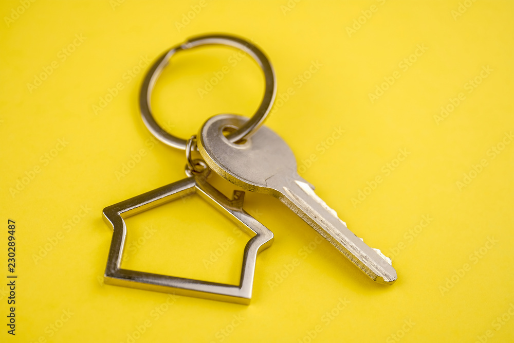 The concept of buying a private house or apartment. Metal keyring in the shape of a house with a metal key on a yellow background. Close up.