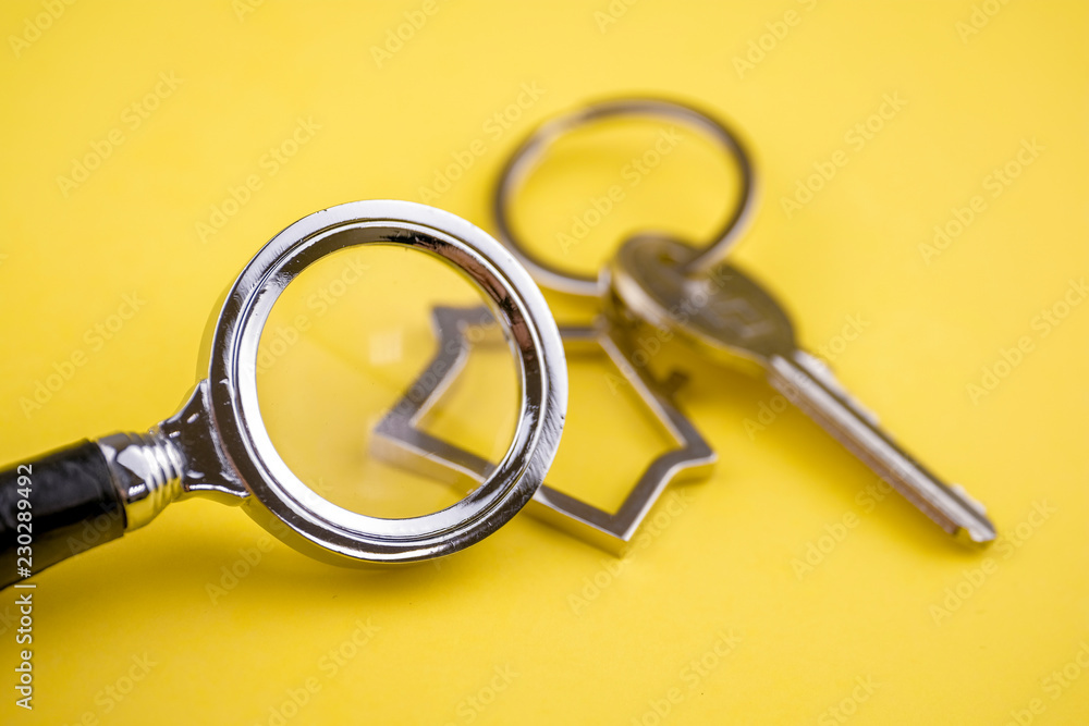 The concept of checking the legal purity of a private house or apartment. Metal keyring in the shape of a house with a metal key under a magnifying glass on a yellow background. Close up. The concept of checking the legal purity of a private house or apartment. Metal keyring in the shape of a house with a metal key under a magnifying glass on a yellow background. Close up.