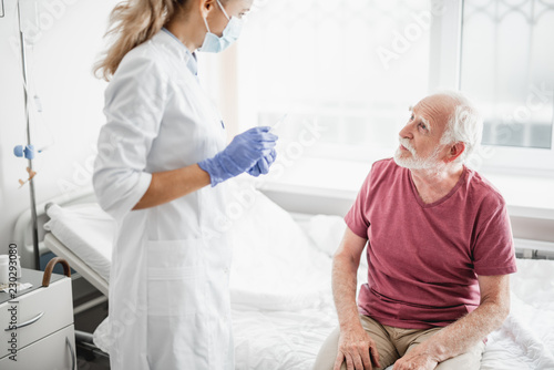 I need to make an injection. Portrait of bearded old man sitting on hospital bed and looking at female medic in sterile blue gloves while she holding syringe