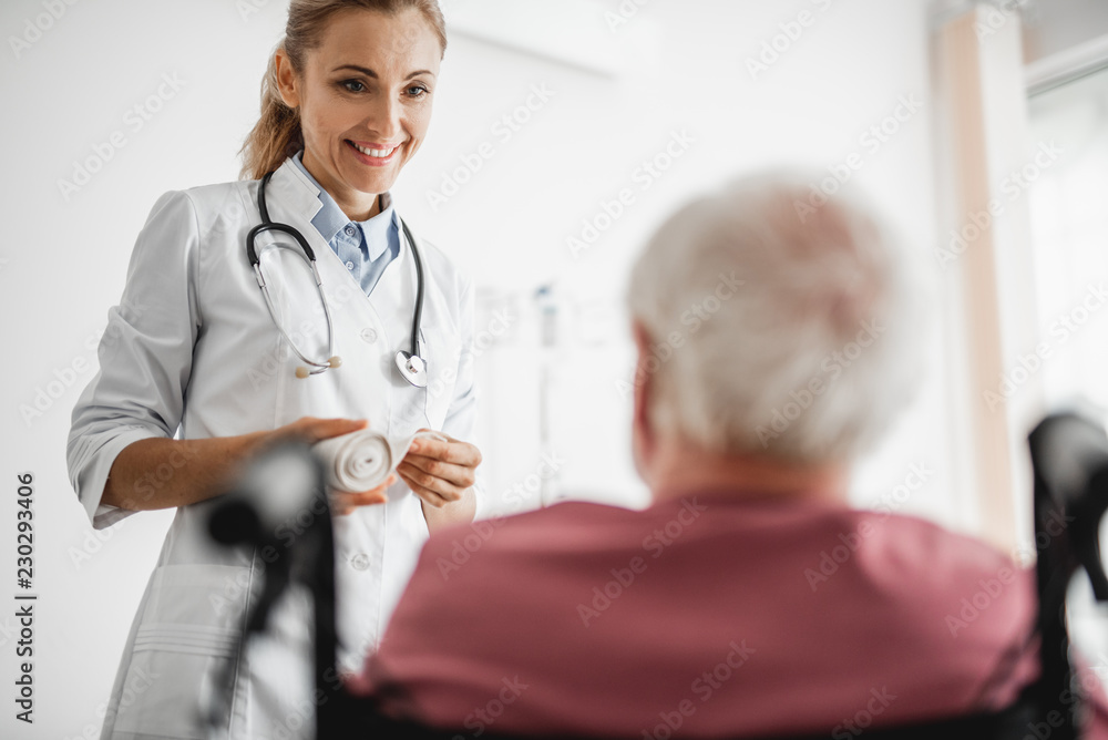 Portrait of smiling lady in white lab coat holding bandage and looking at patient. Focus on young woman