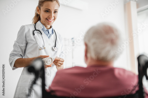 Portrait of smiling lady in white lab coat holding bandage and looking at patient. Focus on young woman