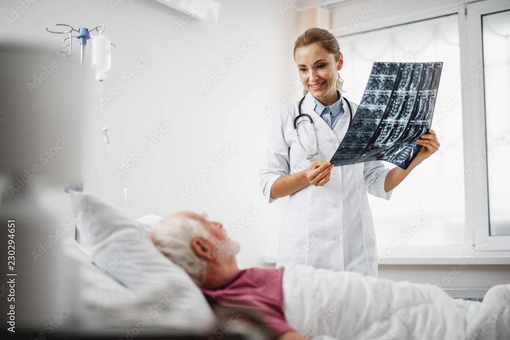 Obraz premium Good news. Portrait of beautiful young lady in white lab coat sharing radiography results with old man. Gentleman lying in hospital bed