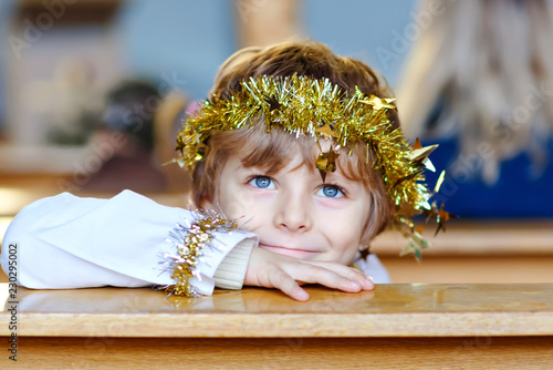 Beautiful little kid boy playing an angel in Christmas story in a church. Happy adorable blond child with lights and xmas tree on background. Celebration of Xmas eve, big christian, catholic holiday. photo