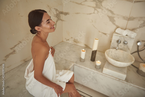 Happy young woman smiling and looking happy while sitting in hammam and touching the towel on her body