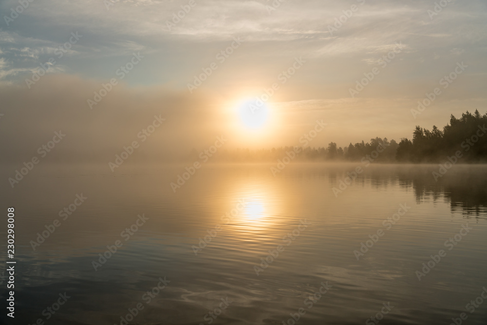 Beautiful golden Sun glows through fog at lake during early sunrise, summer, sky reflection in calm water