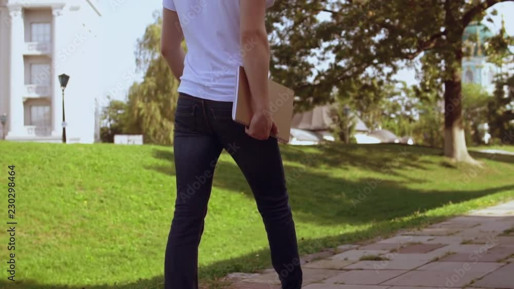 back view man wearing jeans and white t-shirt holding notebook walking on the street in summer city. back view man wearing jeans and white t-shirt holding notebook walking on the street in summer city.