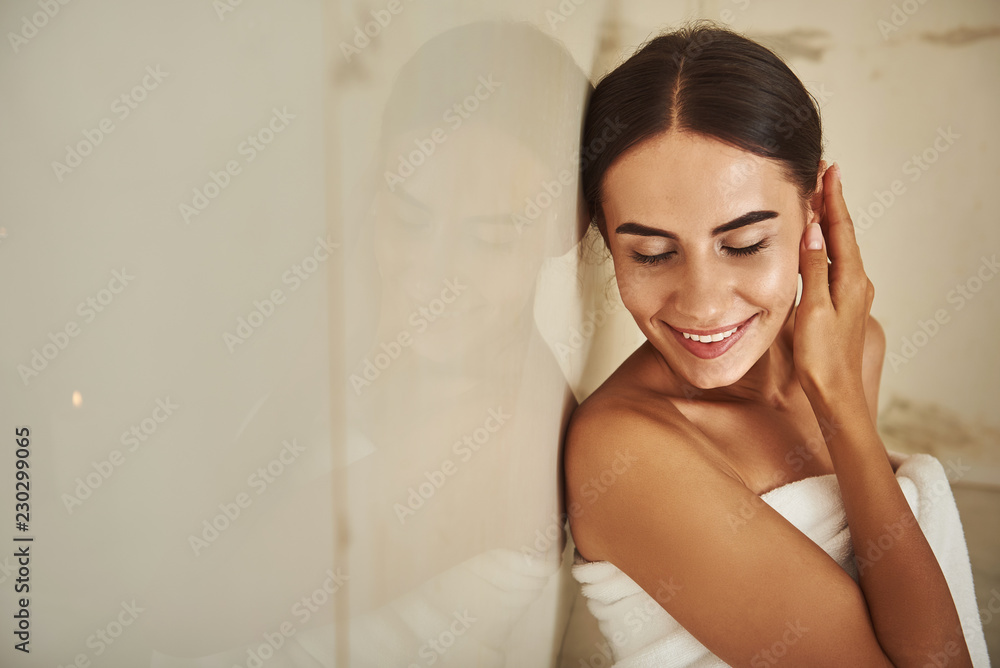 Beautiful dark haired young woman covered in white towel and smiling while touching her ear in hammam Beautiful dark haired young woman covered in white towel and smiling while touching her ear in hammam