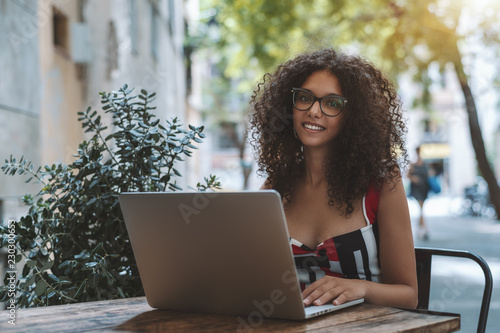 Portrait of beautiful smiling biracial freelancer girl in eyeglasses working via her netbook in an outdoor cafe; a cheerful charming lady with bulky curly hair is using the laptop in a street bar