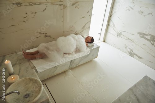 Lots of foam. Peaceful young lady lying in soap foam and smiling while enjoying hammam procedure