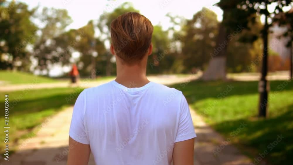 back view caucasian man with blond hair walking in park. guy wearing white t-shirt outdoors slow motion back view caucasian man with blond hair walking in park. guy wearing white t-shirt outdoors slow motion