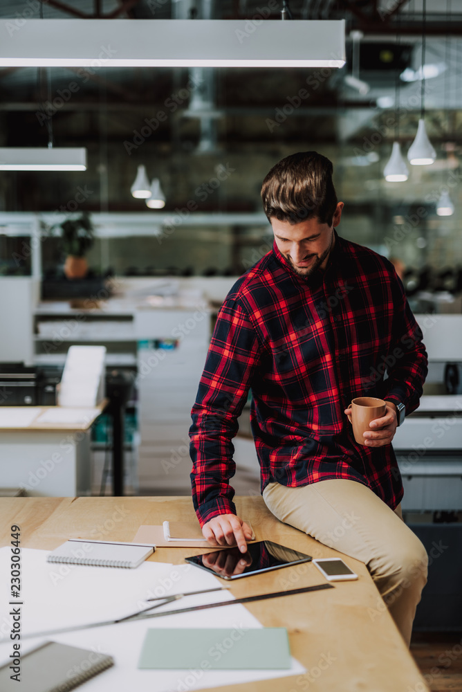 Obraz premium Delighted young bearded man sitting on the table while surfing the internet in the tablet