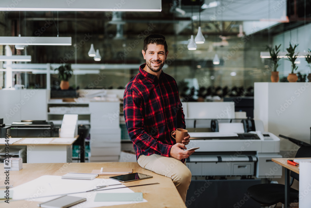 Joyful smiling bearded man using his phone while sitting on the table in his typography office Joyful smiling bearded man using his phone while sitting on the table in his typography office
