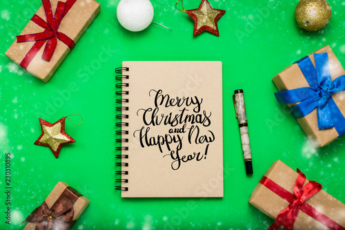 Notebook with with text Merry Christmas and Happy New Year and Christmas-tree decorations, toys and gift boxes on green background. Flat lay, top view