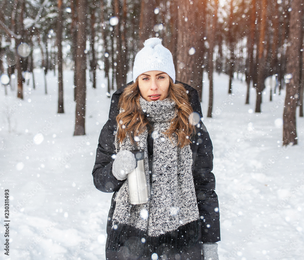 Adorable happy young blonde woman in white knitted hat scarf having fun drinking hot tea from thermos cup snowy winter park forest in nature Adorable happy young blonde woman in white knitted hat scarf having fun drinking hot tea from thermos cup snowy winter park forest in nature
