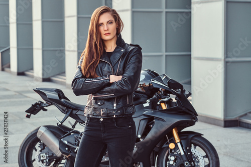 Tela A confident biker girl with her arms crossed next to her superbike outside a building