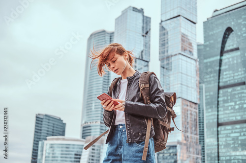 Trendy dressed redhead student girl with tattoos on her face using a smartphone in front of skyscrapers in Moskow city. © Fxquadro