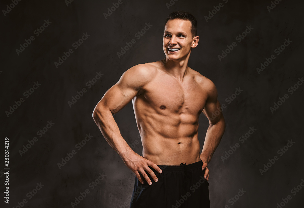 A handsome young fitness model with muscular body posing in studio on dark background.