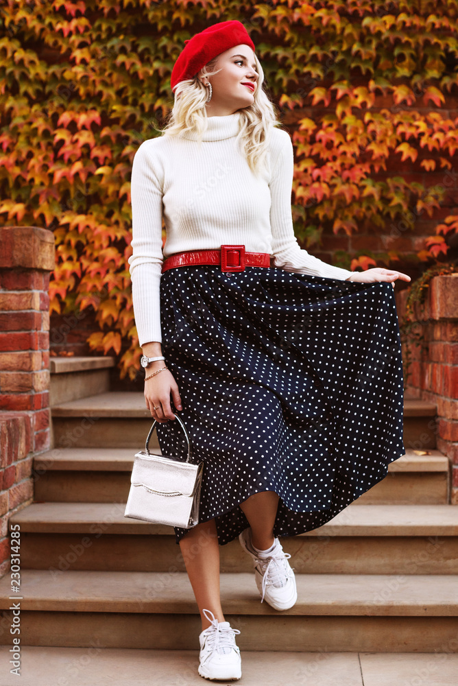 Outdoor full body fashion portrait of young beautiful girl wearing red  beret, belt, white turtleneck, polka dot midi skirt, snickers, wrist watch,  holding small bag, posing in autumn street Stock Photo