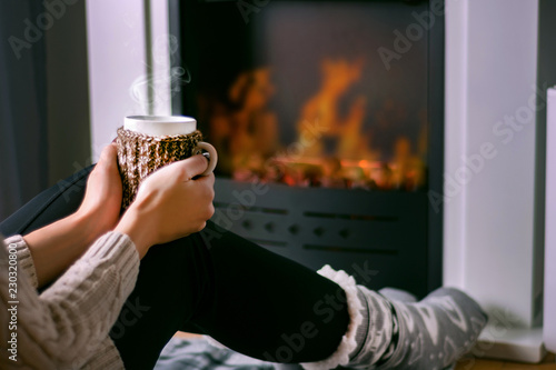 Young girl sitting in front of the fireplace and holding cup of tea in hand on legs and warming. Woman in winter clothes and wool socks. Winter and cold weather concept. Close up, selective focus