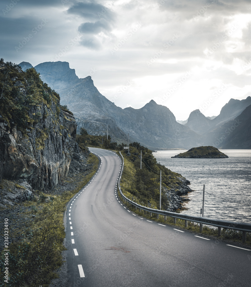 Fototapeta premium Winding road trip towards Henningsvaer on Lofoten Islands with mountains in the background. Cloudy and moody sky with atlantic ocean.
