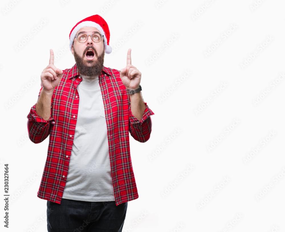 Young caucasian man wearing christmas hat over isolated background amazed and surprised looking up and pointing with fingers and raised arms. Young caucasian man wearing christmas hat over isolated background amazed and surprised looking up and pointing with fingers and raised arms.