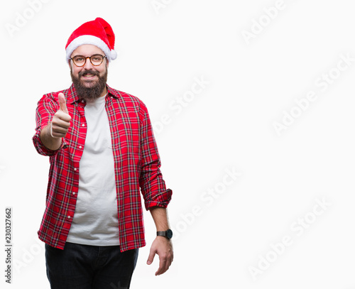 Young caucasian man wearing christmas hat over isolated background doing happy thumbs up gesture with hand. Approving expression looking at the camera with showing success.