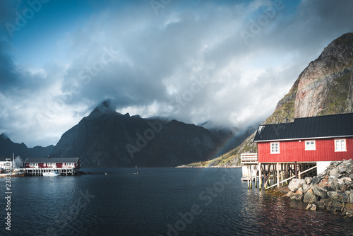 Rainbow ofer red houses rorbuer of Reine in Lofoten, Norway with red rorbu houses, clouds, rainy blue sky and sunny. Bridges and mountains in the background. photo