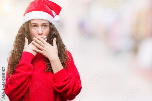 Young brunette girl wearing christmas hat over isolated background shocked covering mouth with hands for mistake. Secret concept.