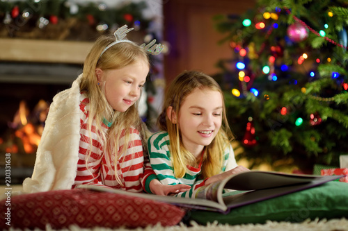 Happy little sisters reading a story book together by a fireplace in a cozy dark living room on Christmas eve.