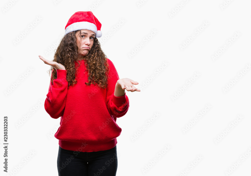 Young brunette girl wearing christmas hat over isolated background clueless and confused expression with arms and hands raised. Doubt concept.