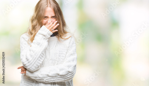 Beautiful young blonde woman wearing winter sweater over isolated background smelling something stinky and disgusting, intolerable smell, holding breath with fingers on nose. Bad smells concept.